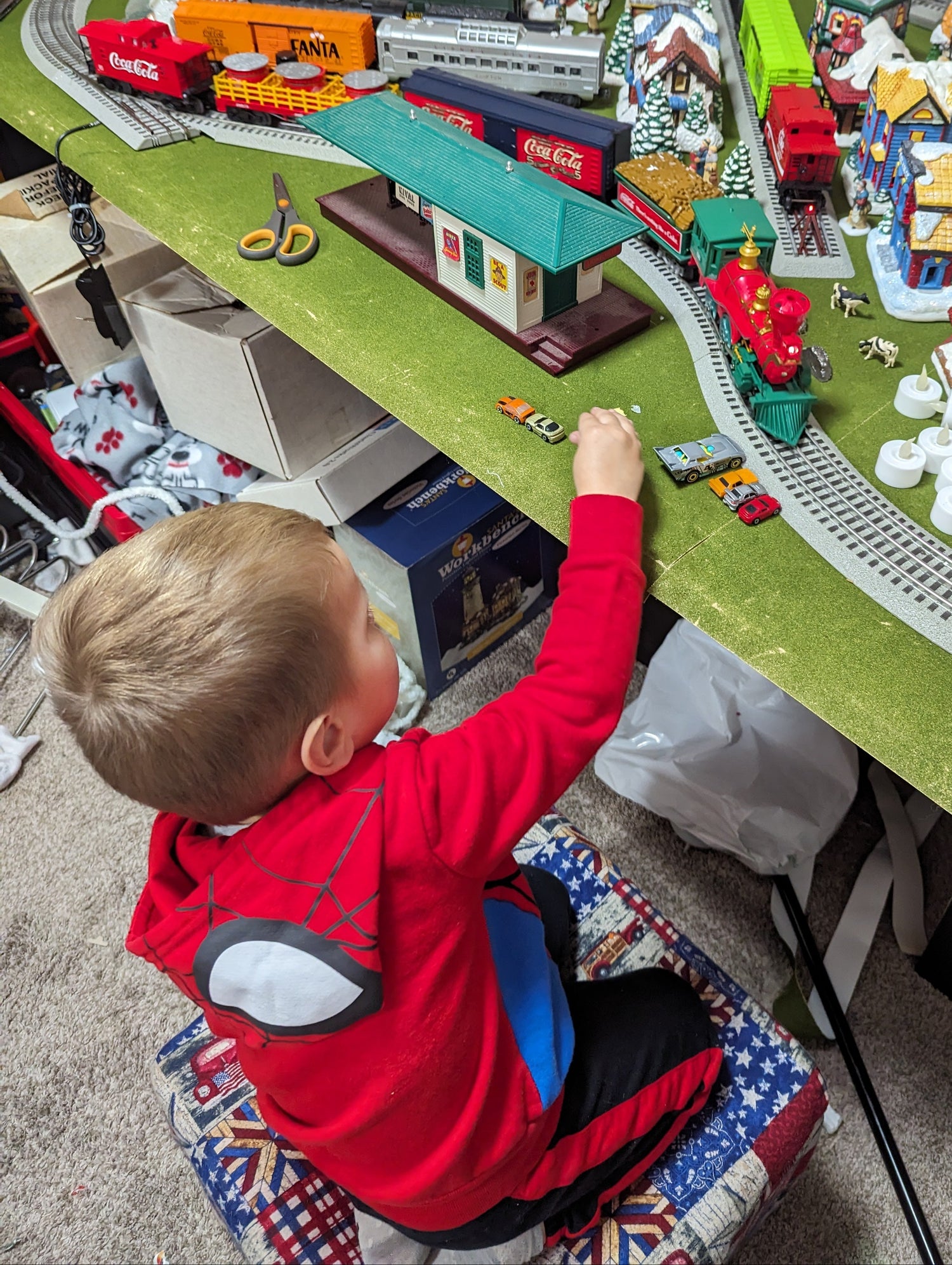 Child at lower left playing with an O gauge holiday train layout, red locomotive and curved track in the upper third.